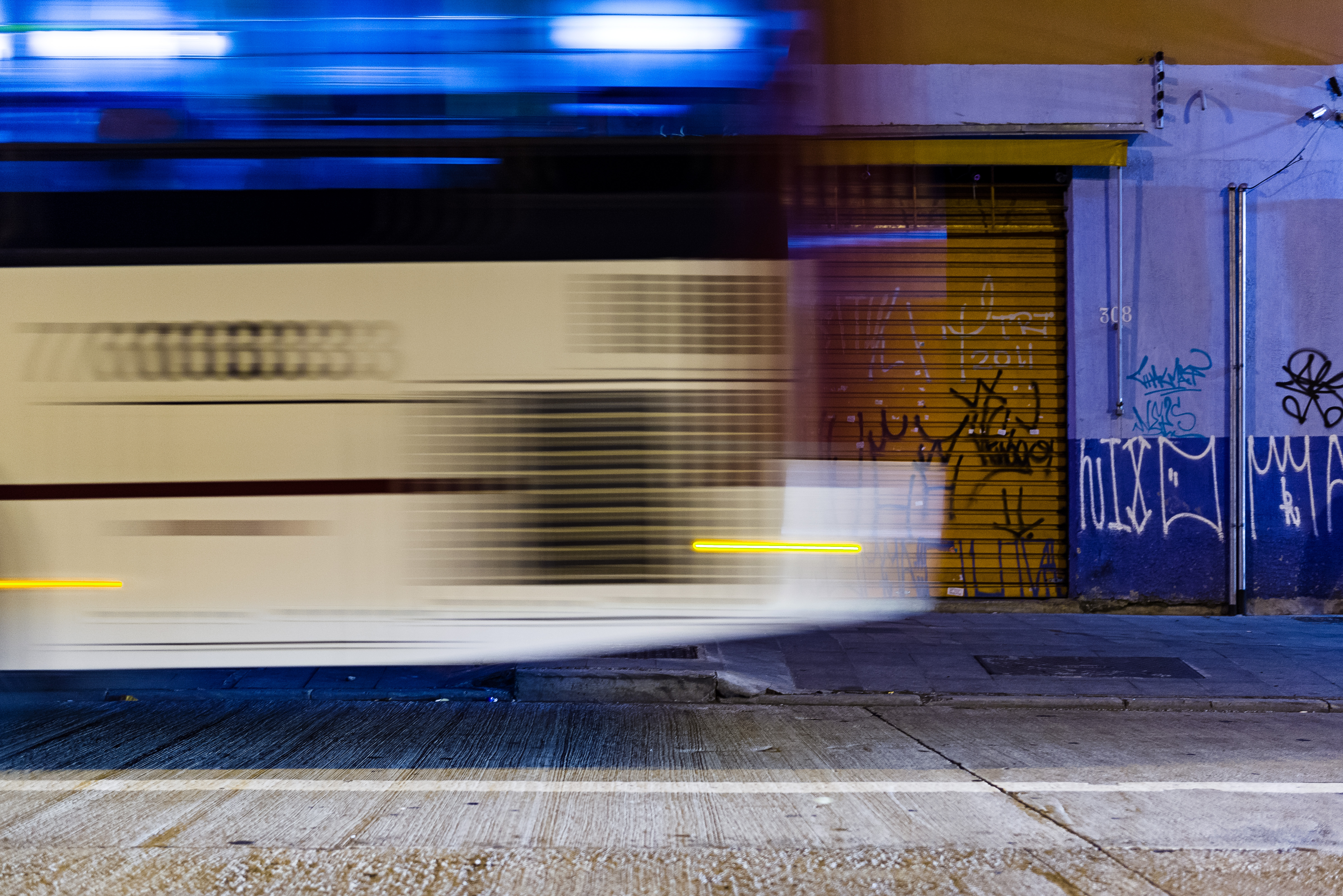 Fotografia colorida tirada ao entardecer de um ônibus em movimento. Atrás do ônibus existe uma porta de alumínio amarela de comércio e a parede do lado metade branca e metade azul com o número 308 atrás de uma barra de ferro que segura um toldo dobrável. Na parede, há duas barras de ferro e, em sua ponta, cabos de energia. Tanto a porta de alumínio quanto a parede ao lado têm inscrições em tinha spray de pixo. A calçada na frente da porta e da parede é limpa e tem um bueiro. Na rua, por onde o ônibus passa, o asfalto possui diversos vincos.