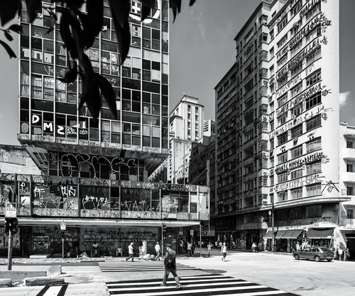 Figura em preto e branco do Largo do Paissandú. À esquerda, um prédio antigo em primeiro plano e, em segundo, logo na parte de cima, algumas folhas de árvores. O Fotógrafo estava embaixo dessa árvore para tirar a foto. Na parte de baixo da foto, duas faixas de pedestres marcadas com linhas brancas horizontais. Ao lado do prédio antigo com pixações, há um prédio ao fundo em branco. Atrás dele, um céu limpo em tom de cinza. Ao lado do prédio branco, ao centro da fotografia, há outro prédio antigo com pixações e coberto em seu lado direito de uma sombra. Do lado esquerdo, já à direita da foto, é iluminada pelo Sol e tem uma cor mais clara, também, cheia de pixações. Abaixo desse prédio, um carro cinza passa pela rua. Nas calçadas, pedestres caminham.