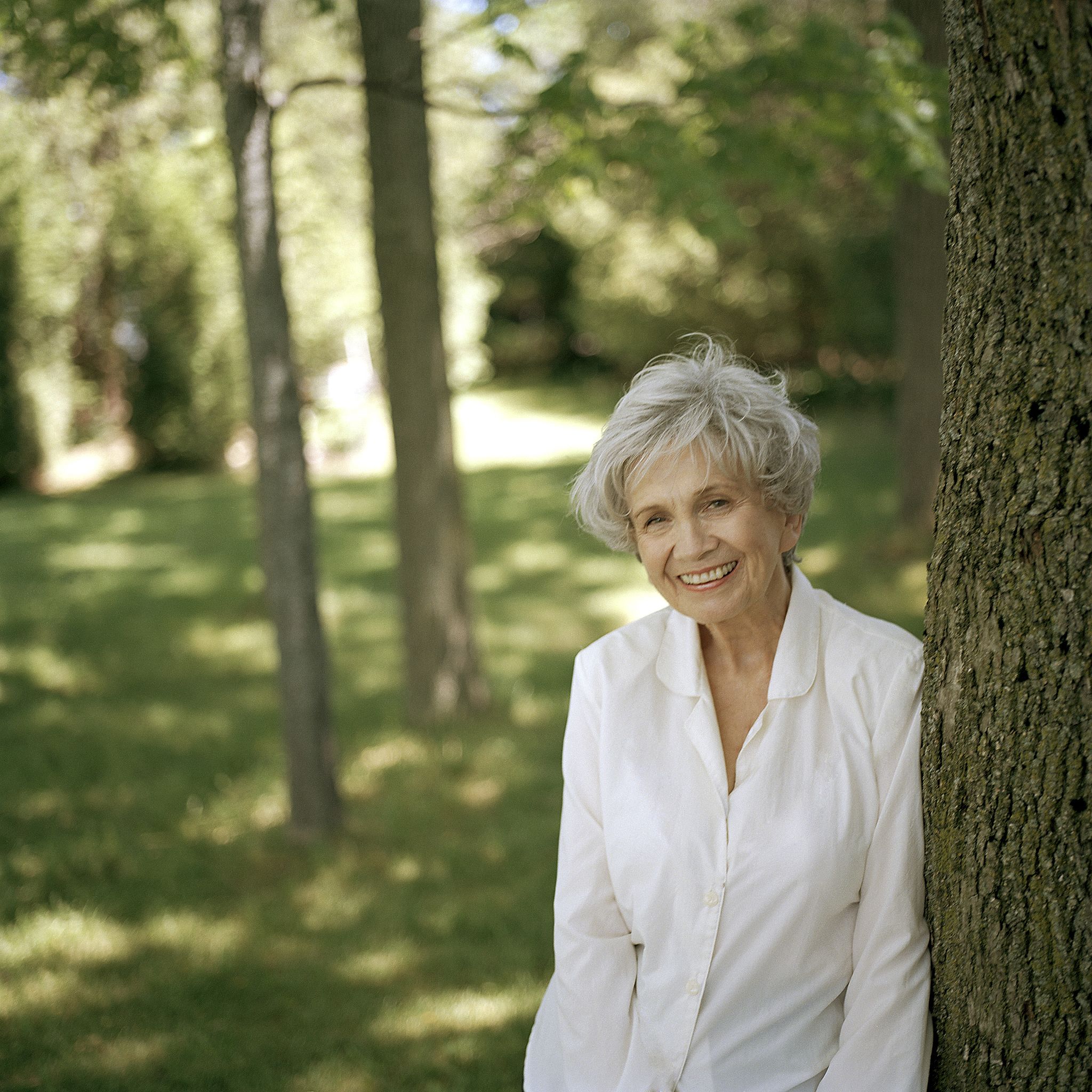 Fotografia de Alice Munro vestida de branco com aproximadamente 70 anos encostada em um árvore em um parque.