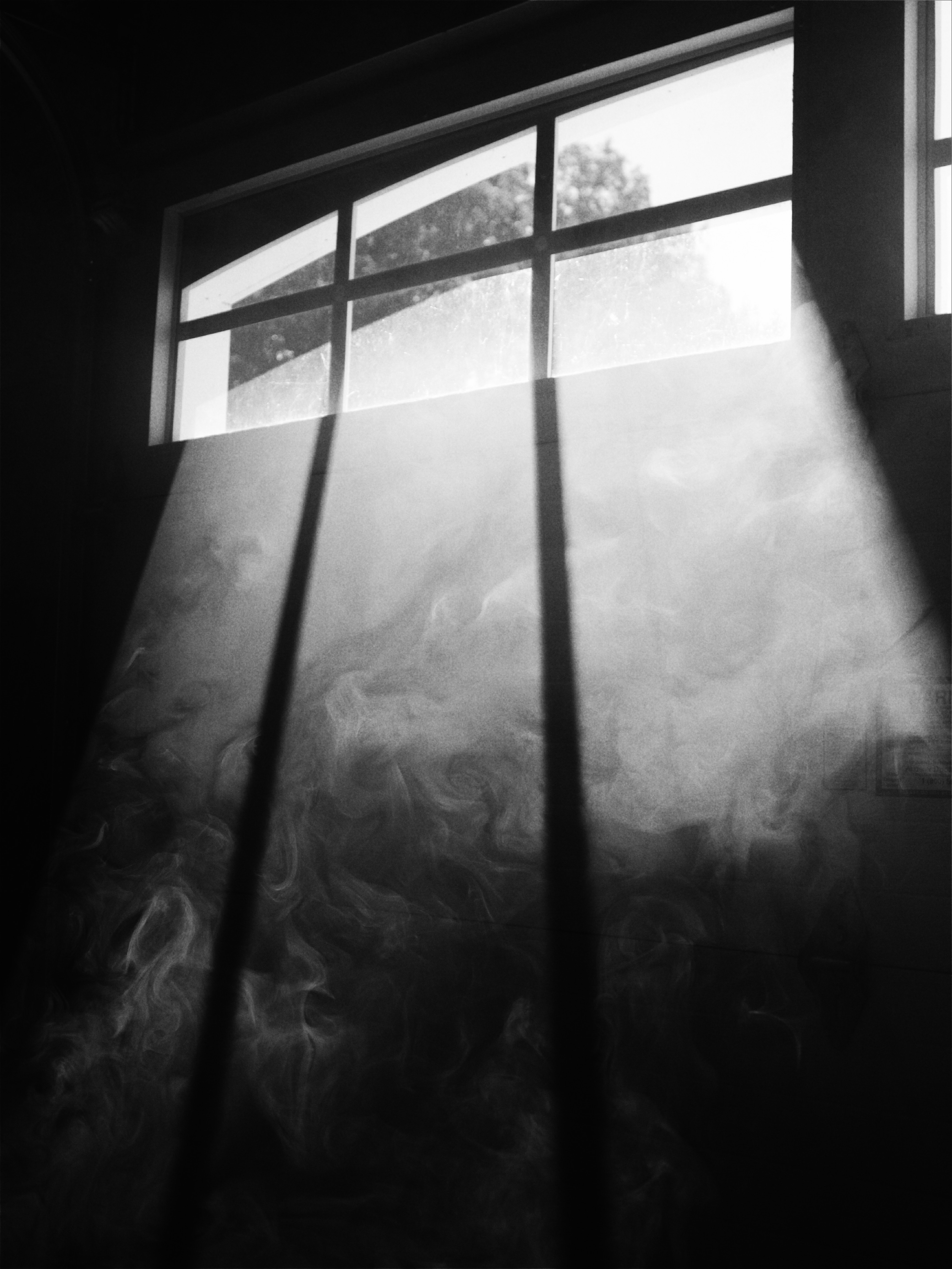 Imagem em preto e branco de uma janela em um espaço que lembra um porão escuro. De fora para dentro uma luz atravessa os vidros da janela e refletem em uma fumaça dentro do espaço.