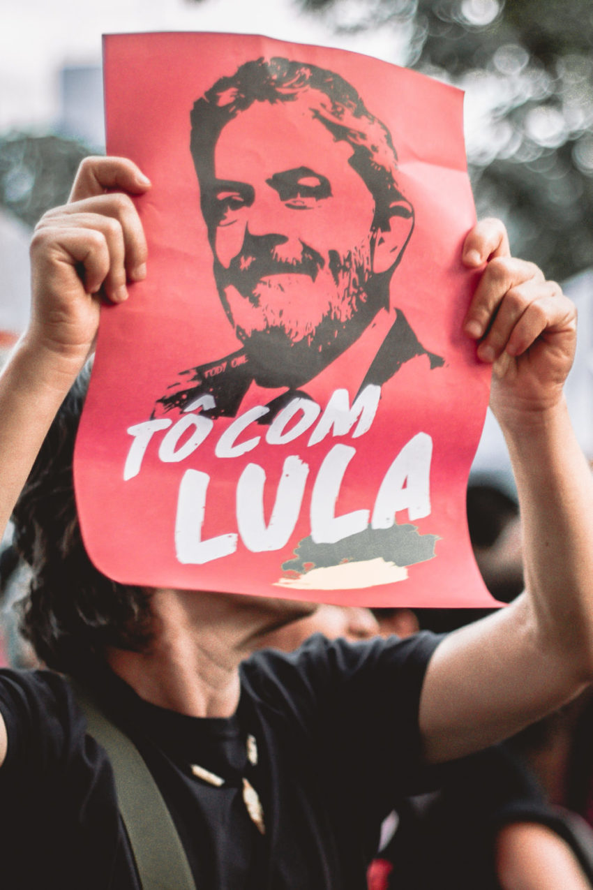 Manifestante segura cartaz vermelho com o desenho do rosto do ex-presidente do Brasil Luiz Inácio Lula da Silva, em preto. Abaixo do desenho do rosto, em branco, os dizeres: "Tô com Lula".