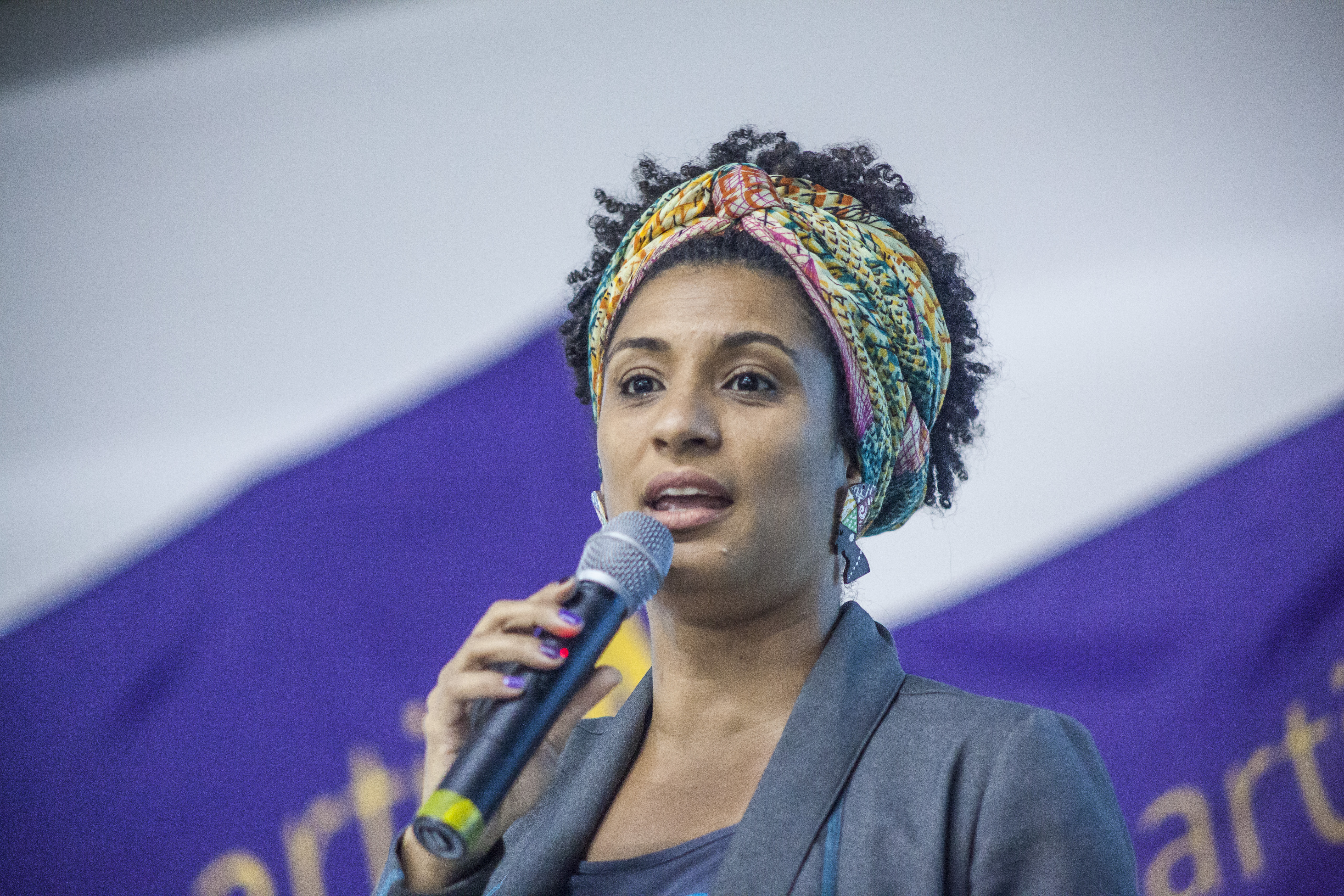 Foto de frente, até a altura do busto, da vereadora carioca Marielle Franco (1979-2018), segurando um microfone durante um discurso.