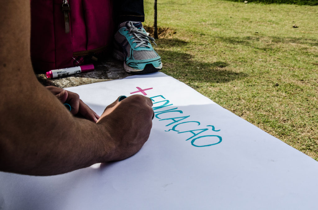 Fotografia de uma mão escrevendo uma mensagem em um cartaz no chão. A mensagem é mais educação.