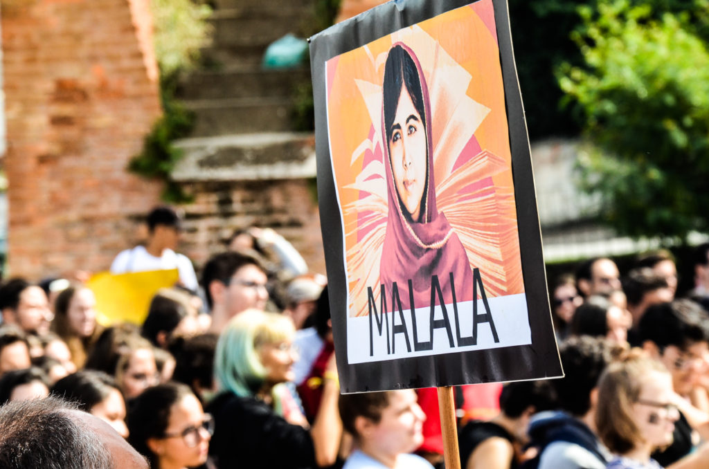 Manifestação com muitas pessoas em segundo plano e em primeiro plano um cartaz com a foto e o nome de Malala.