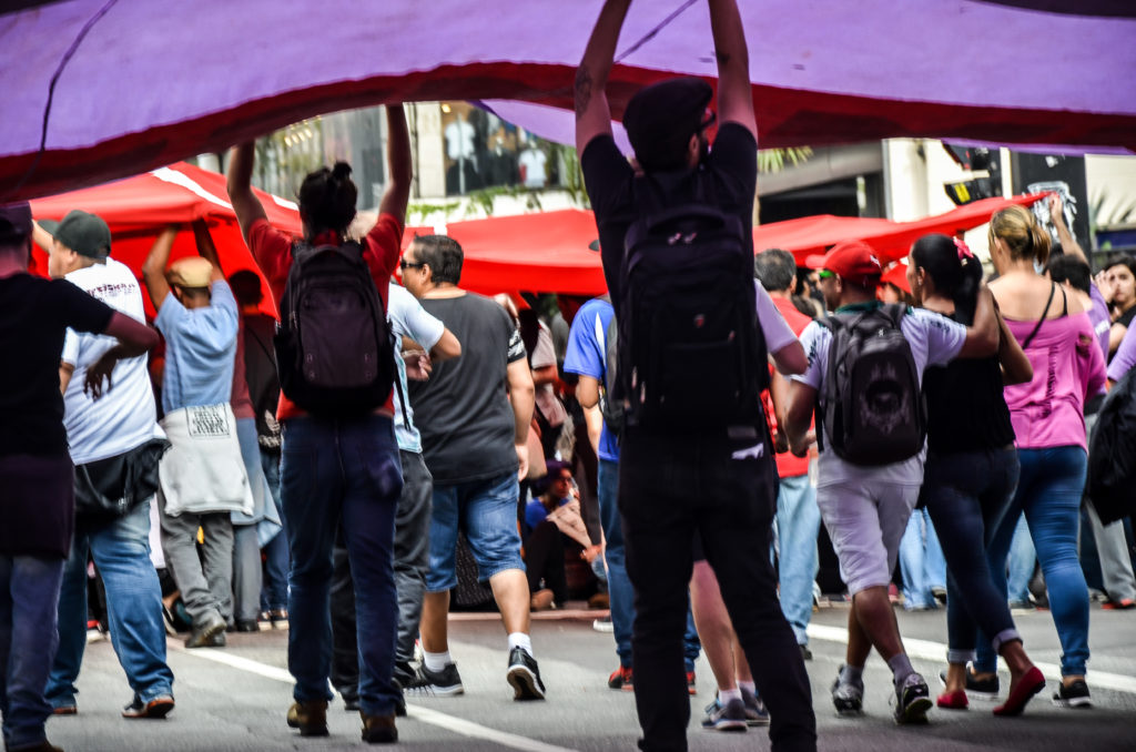 Manifestação em rua com diversos estudantes de costas segurando uma bandeira que os cobre.