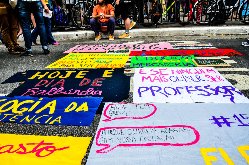 Fotografia de um chão de asfalto em que estão dispostos alguns cartazes com os escritos: Pedagogia da resistência; Educação não é mercadoria; Hoje é dia de Balbúrdia; E se ninguém mais quiser ser professor.