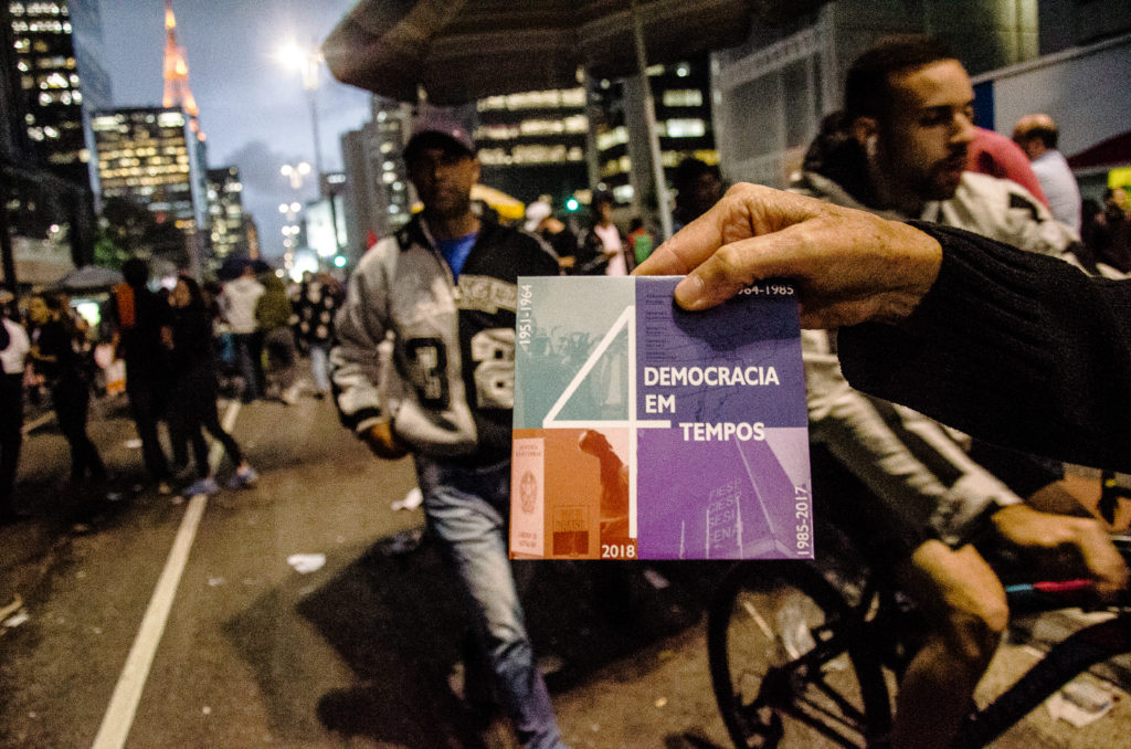 Manifestação na Avenida Paulista com várias pessoas desfocadas e uma mão em primeiro plano segurando um cartaz no qual está escrito Democracia em quatro tempos.