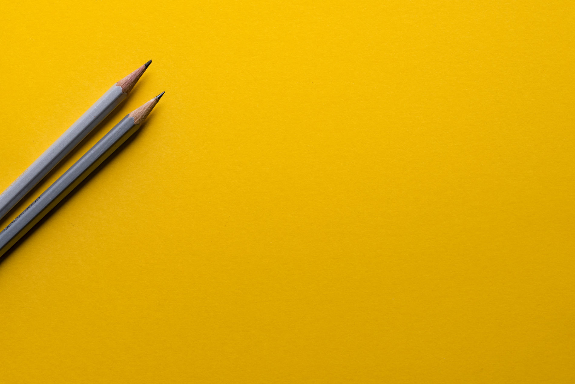 Fundo amarelo com dois lápis cinzas na diagonal do lado esquerdo da foto.