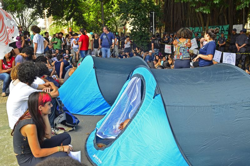 Imagem de um protesto de estudantes na fora da escola estadual Fernão Dias Paes no bairro de Pinheiros em São Paulo. Diversos estudantes em pé e sentados e duas grandes barracas azuis de acampamento na parte centro-direita da imagem. Alguns adultos também aparecem na imagem e também há faixas.
