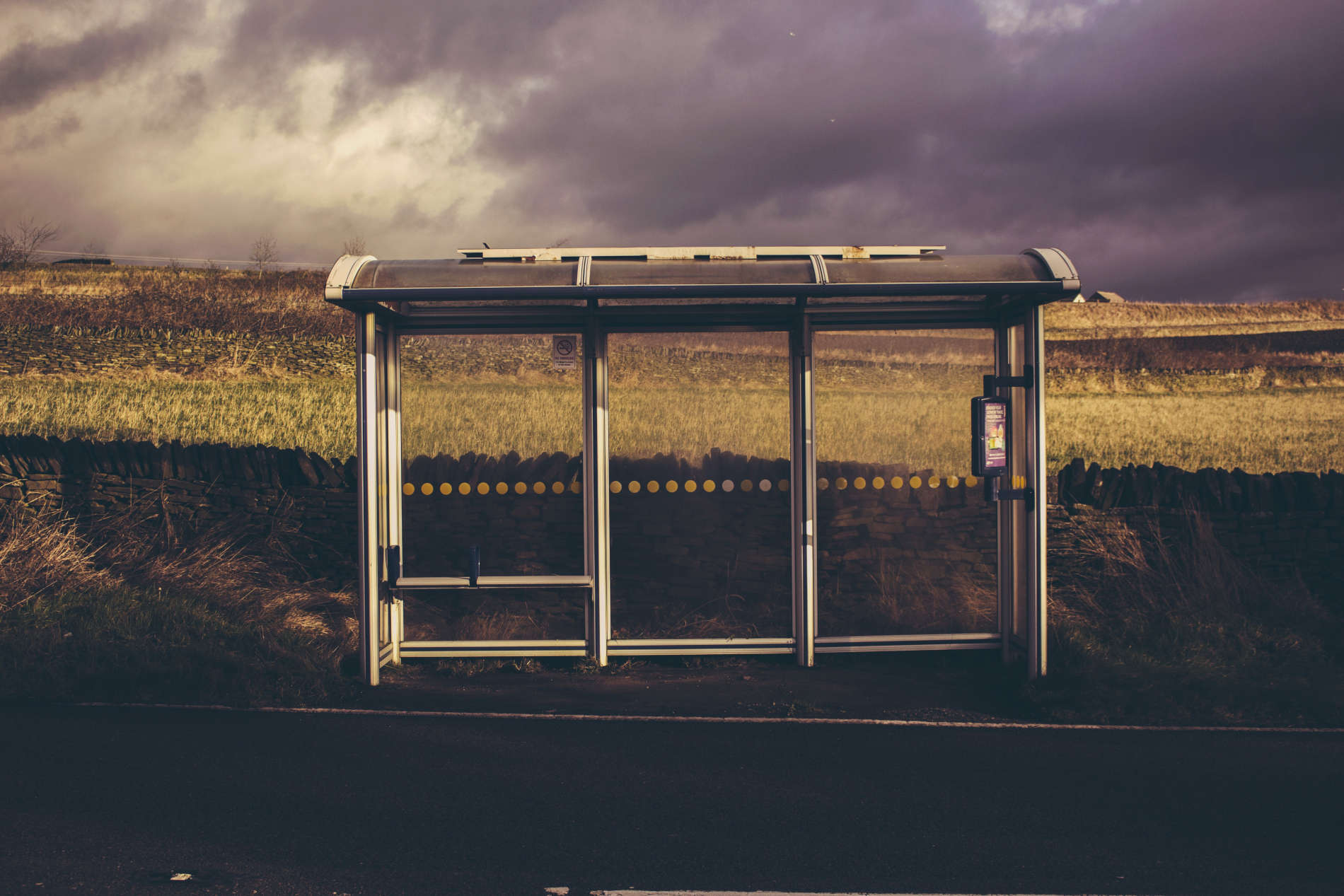 Fotografia de um ponto de ônibus em um campo verde ao fundo. O céu está carregado de nuvens escuras, como se fosse chover a qualquer minuto.