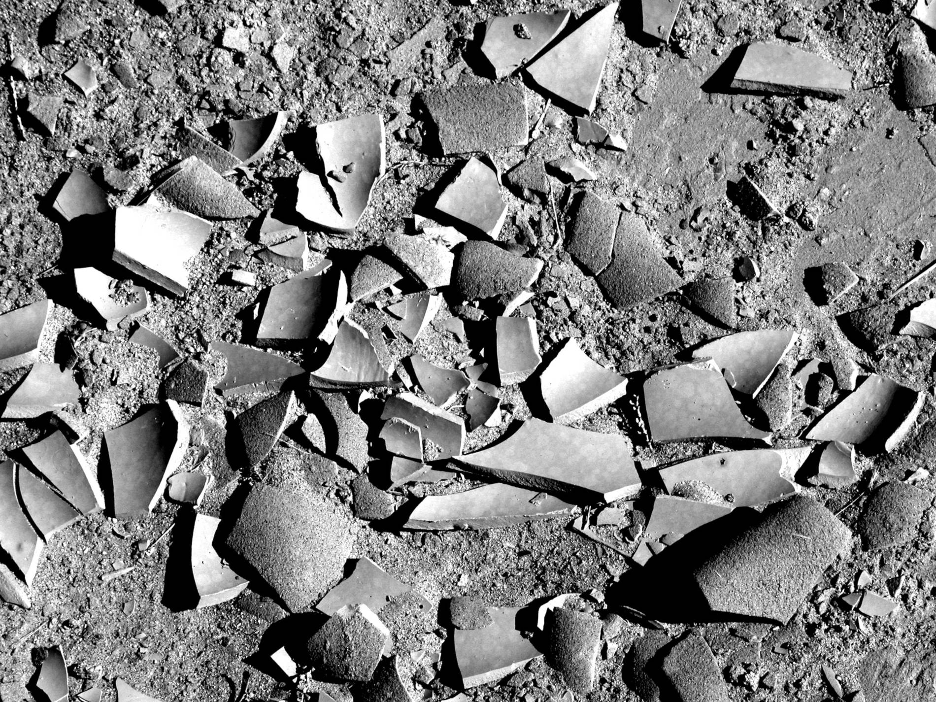 Fotografia em preto e branco de um chão de areia com um utensílio de cerâmica quebrado em várias partes.