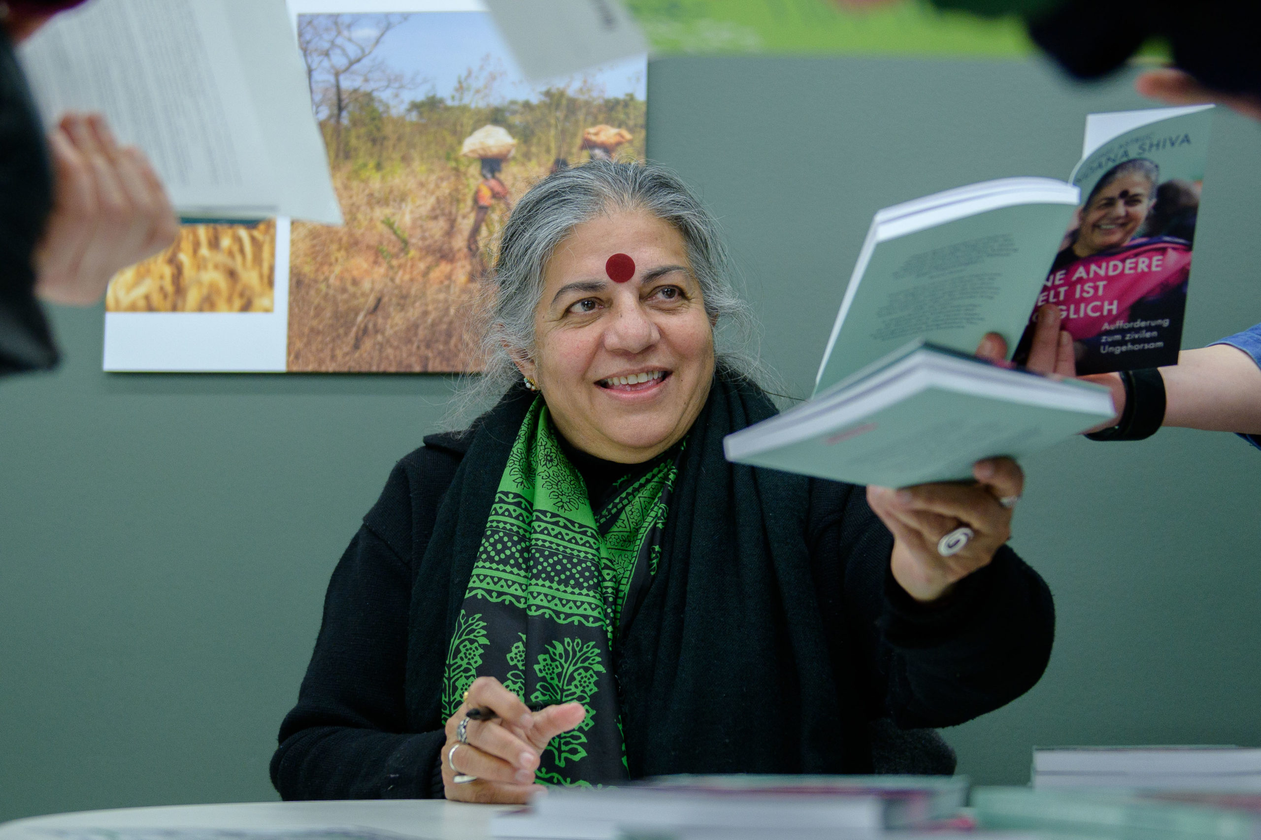 Foto da filósofa Vandana Shiva, vestida de preto com um chale com detalhes verdes, sentada, entregando o seu livro autografado a alguém durante cerimônia de autógrafos, no lançamento do livro.