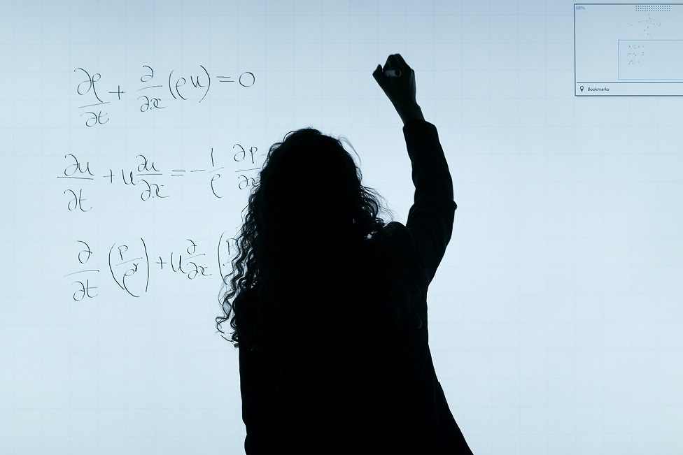 Foto de uma professora de costas, de longos cabelos encaracolados, em pé, em contraluz, escrevendo fórmulas matemáticas em uma lousa branca.