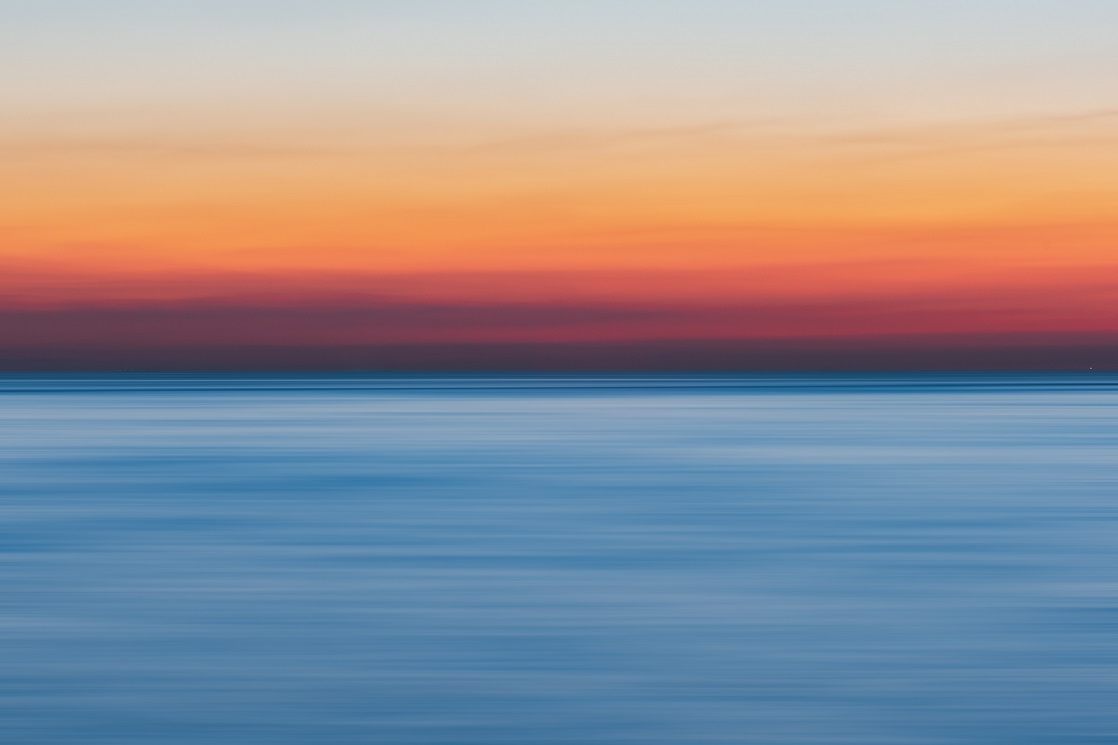 Foto de horizonte no mar aberto, em que a metade superior é um céu em tons rosados, típicos do crepúsculo, e a metade inferior é um mar em tons de azul, de médio para escuro.