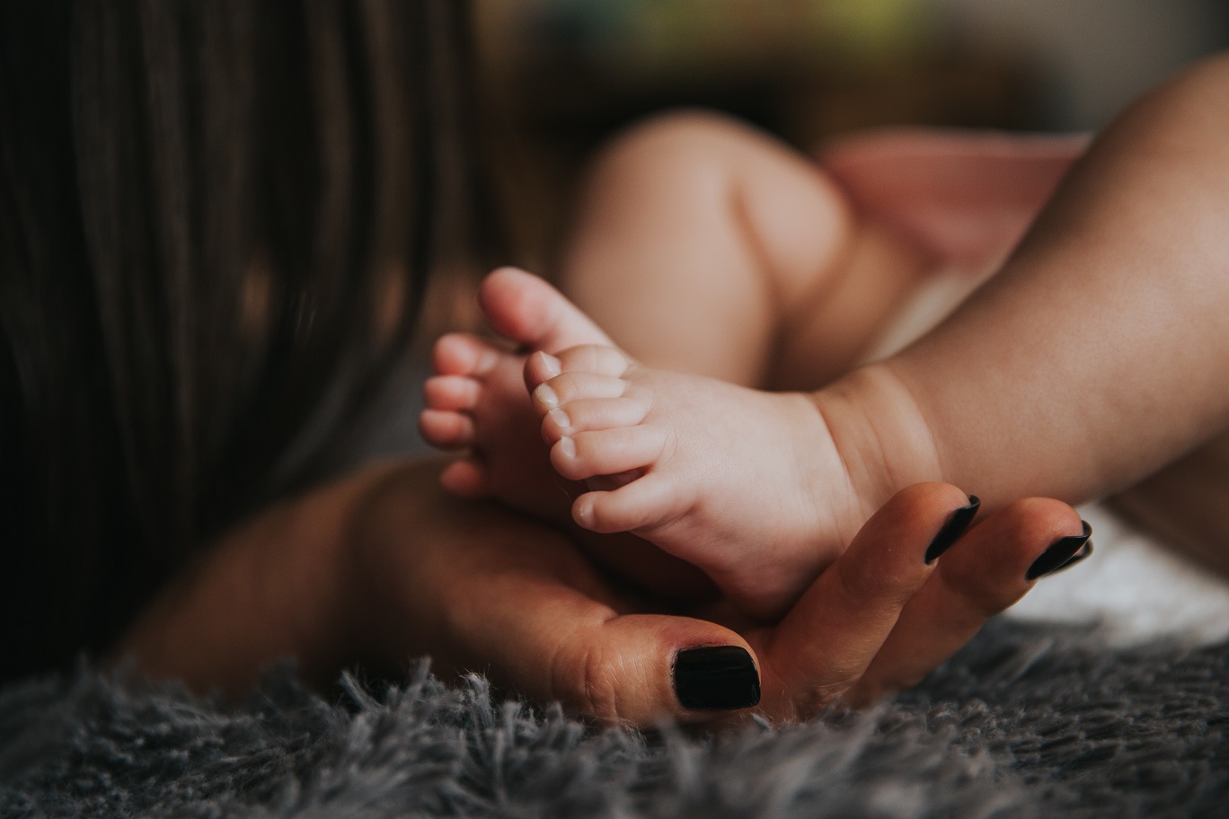 Foto em close de uma mulher adulta, com unhas pintadas de preto, segurando os pequenos pés de uma criança recém-nascida.