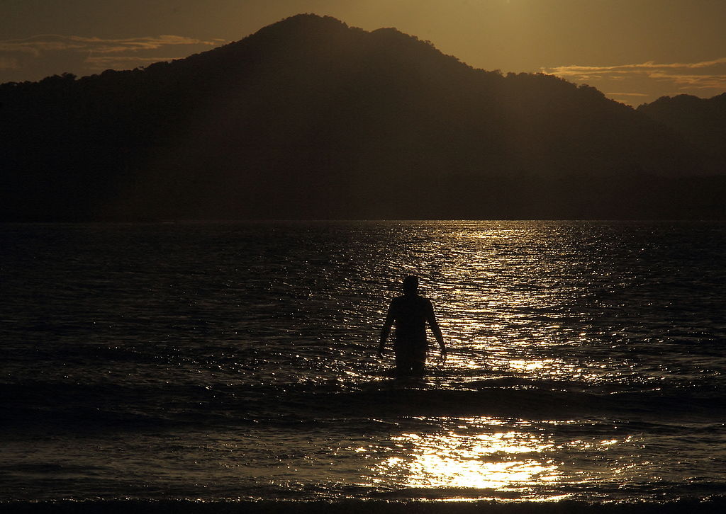 Foto à contraluz de homem caminhando para o mar num fim de tarde na praia.